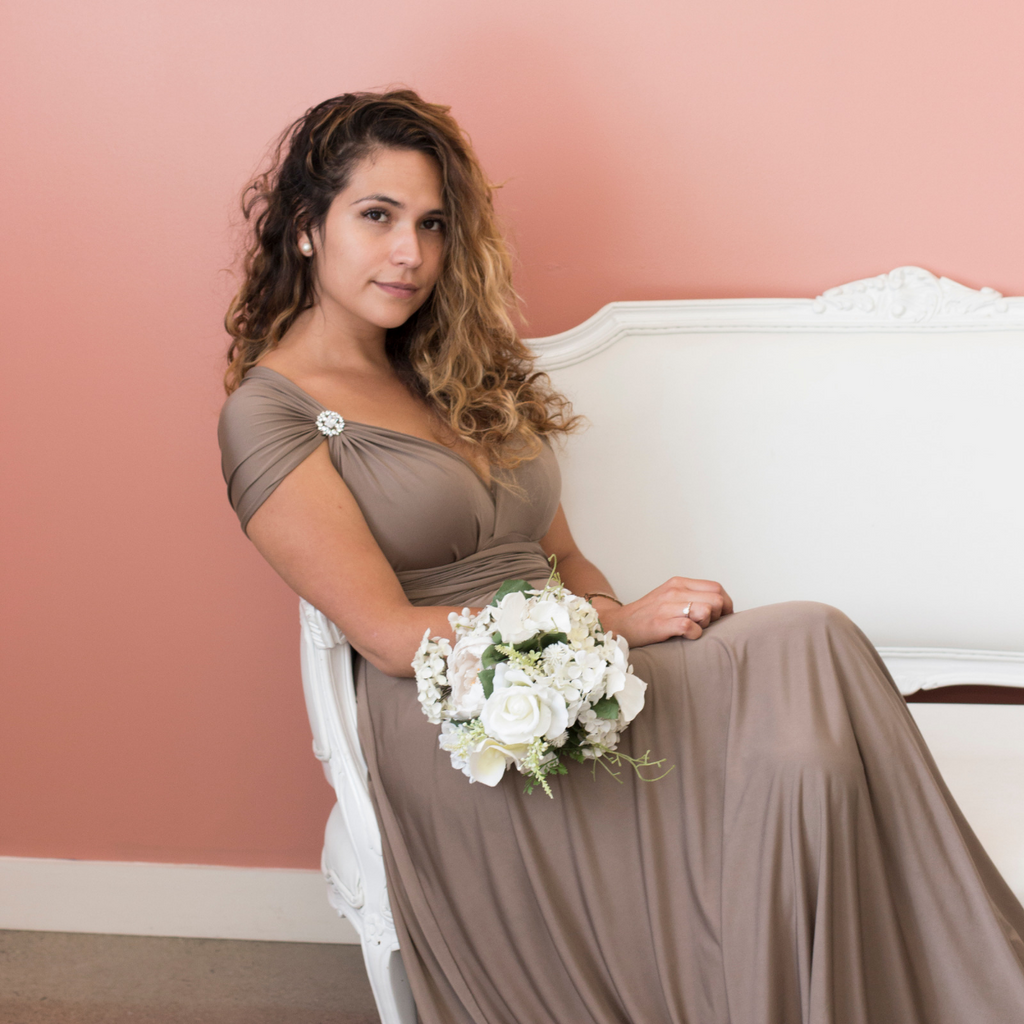 Person wearing Henkaa Sakura Maxi Dress in Truffle Taupe and holding white bouquet while sitting on white settee.