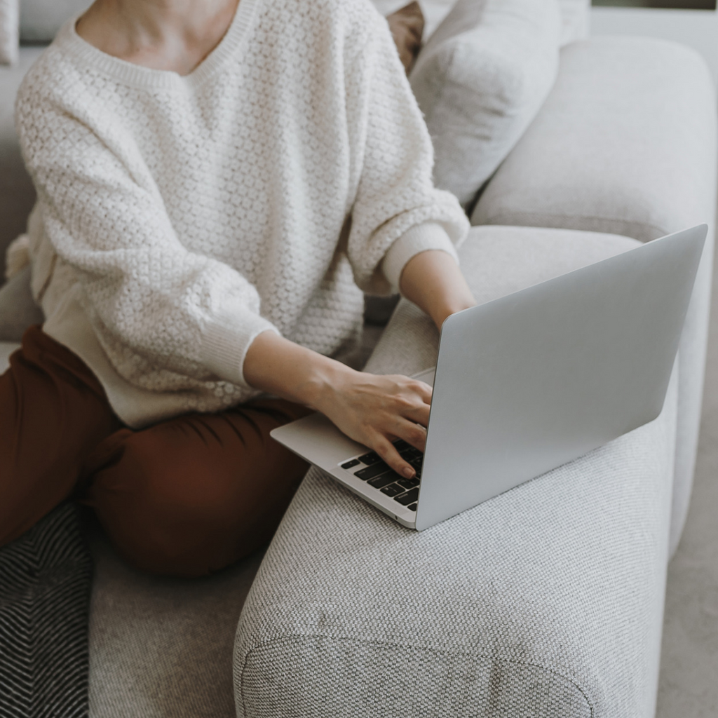 A picture shows a person typing on a slim silver laptop with black keys while seated on a light grey couch in a white sweater and brown pants