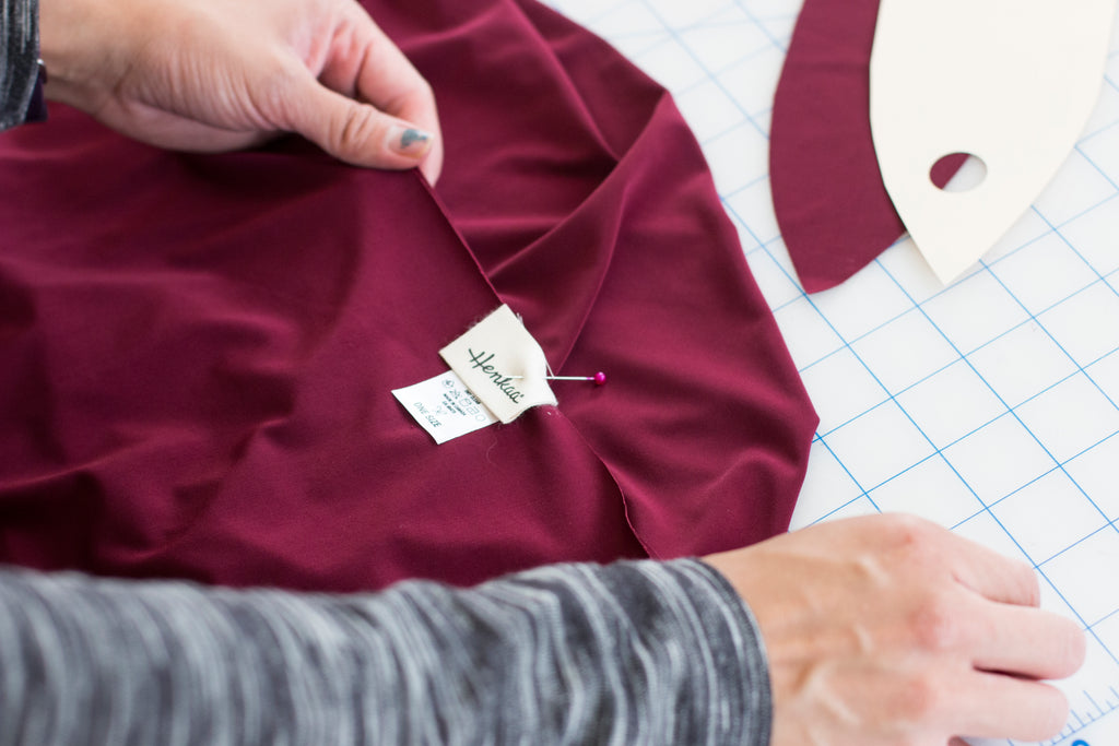A photo shows a person wearing a long-sleeved grey shirt pinning sewing pins through burgundy coloured fabric to attach a Henkaa brand and size label.